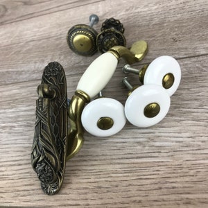 May include: Assortment of cabinet knobs and pulls in a vintage style.  The set includes a white ceramic knob with a gold accent, a gold metal knob with a floral design, and a gold metal pull with a white ceramic handle.