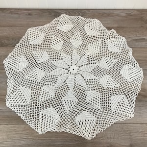 May include: A white crocheted doily with a delicate floral pattern. The doily is round and has a scalloped edge.