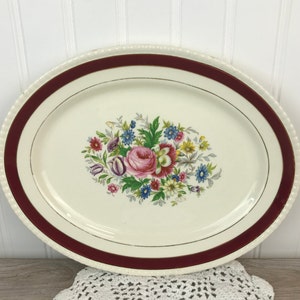 May include: A white oval serving platter with a burgundy rim and a gold trim. The center features a floral design with a pink rose and other colorful flowers.