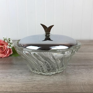 Peut inclure: Un bol en verre transparent avec un motif en spirale et un couvercle argenté avec une poignée décorative. Le bol est sur une surface en bois.