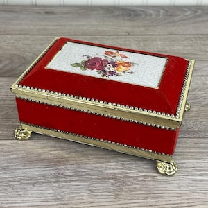 May include: A red velvet jewellery box with a floral design on the lid. The box has gold-coloured trim and decorative feet. The floral design features roses and other flowers.