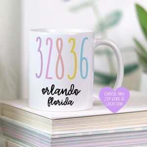 以下が含まれることがあります： カラフルな文字で「32836 orlando florida」と書かれた白いセラミックマグカップ。紫色のハート型のステッカーに「CHOOSE ANY ZIP CODE & LOCATION!」の文字が書かれています。