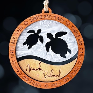 May include: A circular wooden ornament featuring a white marbled resin insert with two black sea turtle silhouettes. The wooden frame is engraved with the text "TURTLE BAY, O'AHU" and the names "Amanda + Richard" are engraved on the lower wood section.