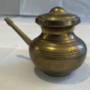 May include: A small, antique brass pot with a lid and a spout. The pot has a rounded body with horizontal ridges and a small, round lid. The spout extends from the side. The brass has a warm, golden color.