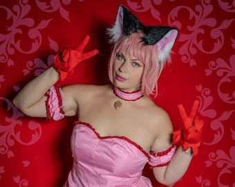 May include: A woman wearing a pink and white dress with a white lace trim. She is wearing a black cat ear headband and red gloves. She is posing with a peace sign in each hand. The background is a red and white patterned wallpaper.
