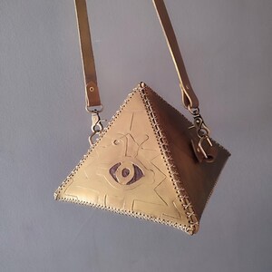 Op de afbeelding: Een piramidevormige crossbodytas van bruin leer met stiksels en een schouderriem. De tas heeft een reliëf oogsymbool en een decoratief patroon. Het heeft een metalen sluiting en hardware.