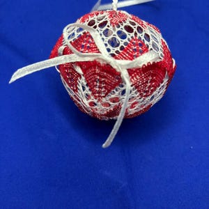 May include: A round, decorative ornament with a red and white lace design. The ornament is spherical and has a white ribbon for hanging. The lace pattern is intricate, with a mix of solid and open areas. The background is a solid blue.