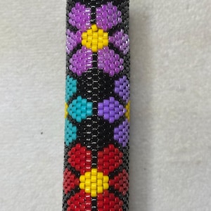 Puede incluir: Un objeto cilíndrico negro con cuentas con un patrón floral. Las flores son en tonos de morado, rojo, amarillo y turquesa. Las cuentas están tejidas apretadamente, creando una superficie texturizada. El objeto es probablemente un estuche para encendedor o un accesorio similar.