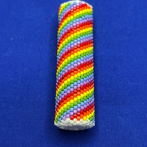 Könnte beinhalten: Ein zylindrisches Objekt, wahrscheinlich eine Feuerzeugabdeckung, ist aufwendig mit kleinen, farbenfrohen Perlen gewebt. Das Design zeigt ein lebendiges Regenbogenmuster aus Rot, Orange, Gelb, Grün, Blau und Lila. Die Basis ist weiß, der Hintergrund ist einfarbig blau.