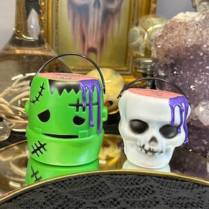 Könnte beinhalten: Zwei Halloween-Süßigkeiteneimer, einer grün in Form von Frankenstein und einer weiß in Schädelform, mit lila tropfender Farbe. Sie haben schwarze Griffe und stehen auf einem goldfarbenen Tablett. Im Hintergrund befindet sich ein Totenkopf-Gemälde.