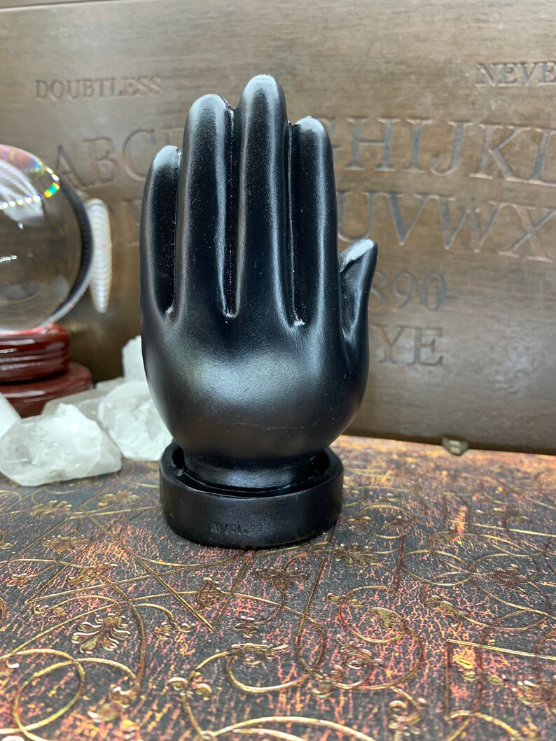 Peut inclure: Une sculpture de main noire stylis&eacute;e avec une paume ouverte. La sculpture est pos&eacute;e sur une base ronde et repose sur une surface d&eacute;corative. L'arri&egrave;re-plan comprend une boule de cristal, des cristaux et une planche de bois avec des lettres et des chiffres.