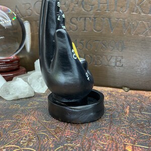 Peut inclure: Un bougeoir noir en forme de main avec des symboles blancs et un accent jaune. Le bougeoir est sur une base ronde. L'arri&egrave;re-plan comprend un tableau Ouija en bois et une boule de cristal.