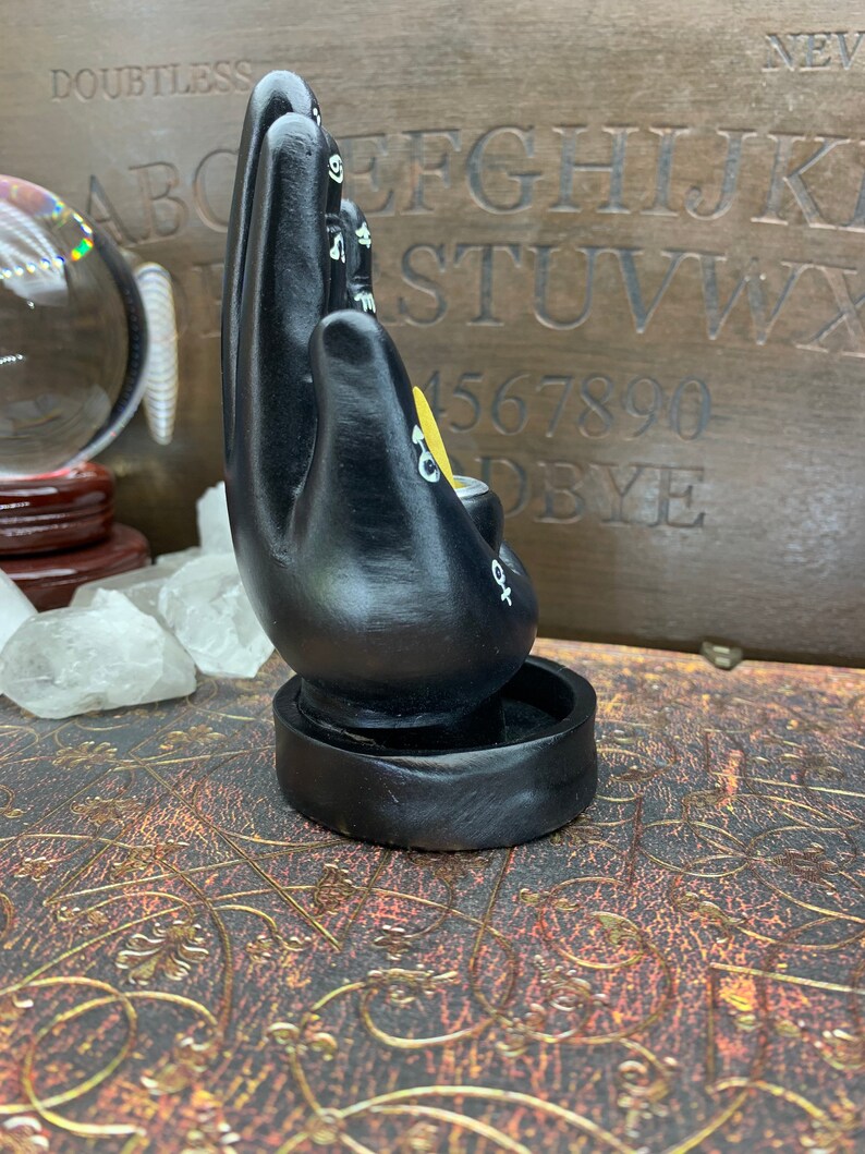 Peut inclure: Un bougeoir noir en forme de main avec des symboles blancs et un accent jaune. Le bougeoir est sur une base ronde. L'arri&egrave;re-plan comprend un tableau Ouija en bois et une boule de cristal.