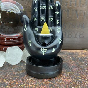 Peut inclure: Porte-encens noir en forme de main avec des symboles astrologiques et un c&ocirc;ne d'encens dor&eacute;. Le support a une base circulaire et un petit r&eacute;ceptacle pour recueillir les cendres. Une boule de cristal et des cristaux sont en arri&egrave;re-plan.
