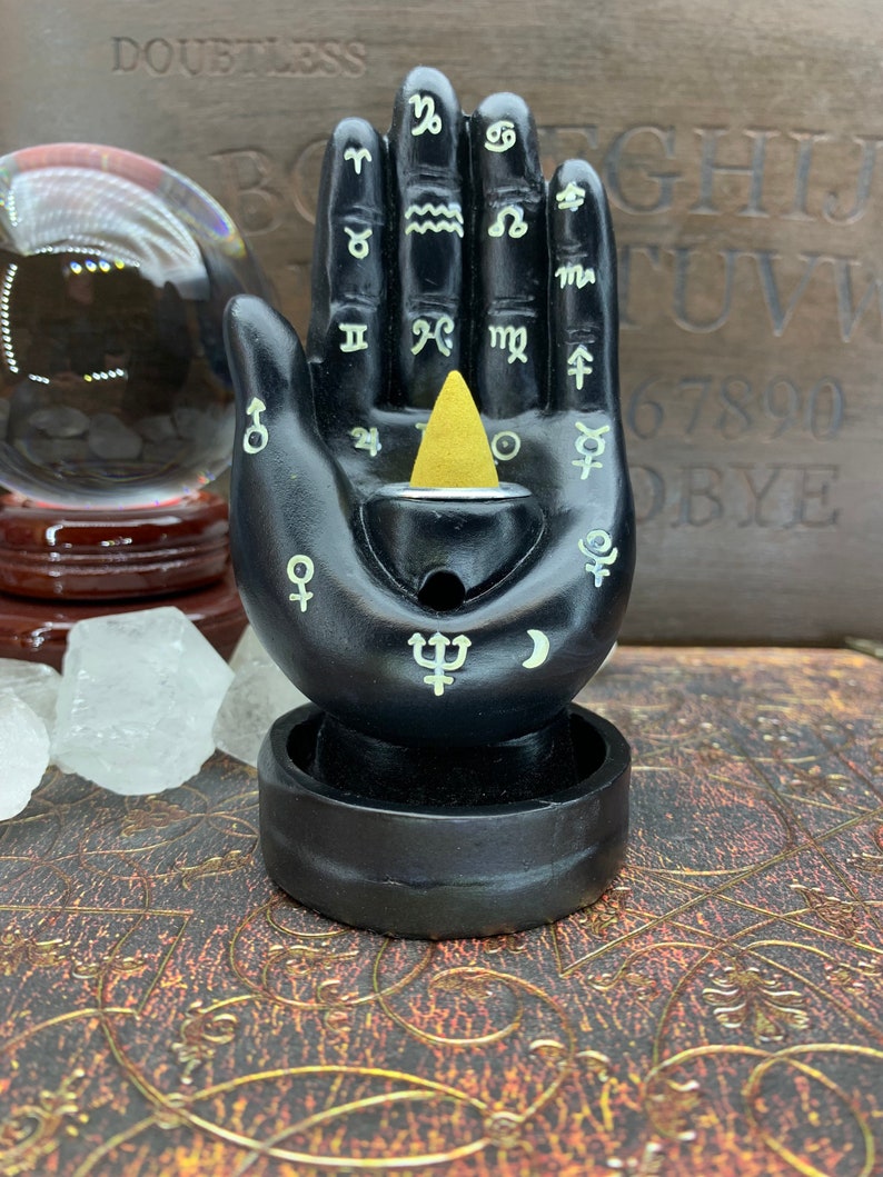 Peut inclure: Porte-encens noir en forme de main avec des symboles astrologiques et un c&ocirc;ne d'encens dor&eacute;. Le support a une base circulaire et un petit r&eacute;ceptacle pour recueillir les cendres. Une boule de cristal et des cristaux sont en arri&egrave;re-plan.