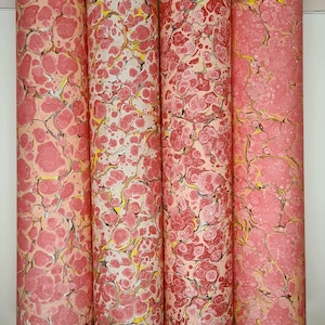 Könnte beinhalten: Vier Rollen Dekorpapier mit Marmormuster. Das Papier weist eine Mischung aus rosa, rot, gelb und weißen Farben auf. Die Rollen stehen aufrecht vor einem weißen Hintergrund. Das Papier hat ein einzigartiges, künstlerisches Design.