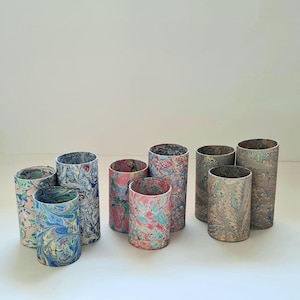 May include: A selection of cylindrical decorative containers with marbled patterns in various colours. The containers vary in height and diameter, with patterns including blue, pink, green, and grey swirls. The containers are arranged on a white surface.