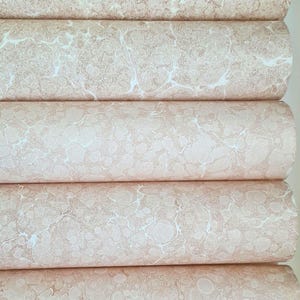 May include: Five cylindrical rolls of paper stacked horizontally, each featuring a marbled pattern with soft beige and light brown swirling textures against a creamy white background, creating a continuous visual effect across the stacked surfaces.