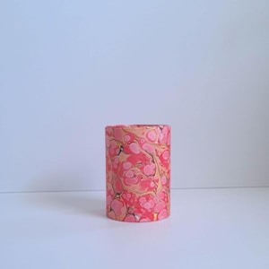 May include: A cylindrical container with a marbled pattern in shades of pink, red, and gold. The container is a solid color and stands upright against a white background. The design is reminiscent of decorative paper.