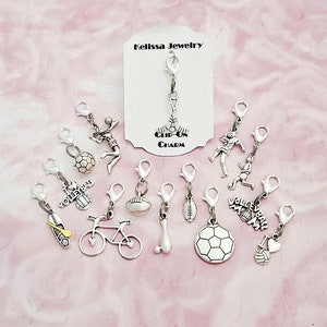 May include: A collection of silver-toned clip-on charms featuring various sports themes, including a volleyball, football, baseball, soccer ball, basketball, bicycle, bowling pin, and a hockey stick. The charms are displayed on a pink surface with a white card that reads "Melissa Jewelry Clip-On Charm".