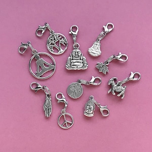 May include: A collection of silver charm pendants with various designs, including a peace sign, a hand, a tree, a meditating figure, a goat, and a Buddha. The charms are on a pink background.