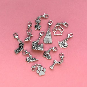 May include: A collection of silver charm pendants featuring various animal designs, including cats, dogs, and a paw print. The charms are arranged on a pink background.