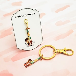 May include: A gold-colored key chain with a giraffe charm. The giraffe is patterned with green, orange, and brown. The charm is attached to a gold-colored ring and a lobster clasp. The text "Clip on Charm" is visible on a white card behind the charm.