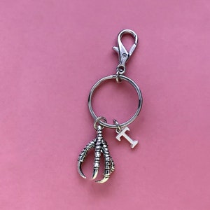 May include: Silver key chain with a claw charm and a letter 'T' charm. The key chain is on a pink background.