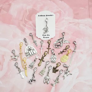 May include: A collection of silver-toned clip-on charms with various designs, including swords, crosses, a dragon, a unicorn, and a knight's helmet. The charms are displayed on a pink floral background. The text "Bellisa Jewelry" and "Clip-On Charm" are visible.