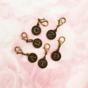 May include: Six gold-toned metal stitch markers with letter charms. The letters are C, N, L, A, T, and D.