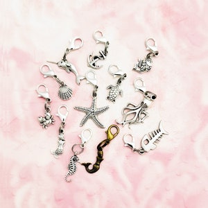May include: A set of 12 silver-toned metal charms featuring various sea creatures, including a starfish, a seahorse, a mermaid, a crab, a turtle, a fish, an octopus, a seashell, an anchor, and a narwhal. The charms are attached to lobster clasps, making them ideal for adding a touch of whimsy to crafts, jewelry, or bags.
