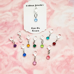 May include: Assortment of clip-on charms with various colored rhinestones. The charms are silver-toned and feature a lobster claw clasp. The charms are displayed on a pink background with a white card that reads "Melissa Jewelry Clip-On Charm".