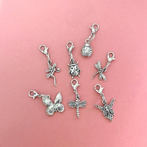 May include: A collection of silver-toned charms featuring various designs, including a fairy, butterfly, ladybug, and dragonfly. Each charm is attached to a clasp, perfect for jewelry making or crafting projects. The charms are displayed on a pink background.