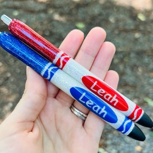 K&ouml;nnte beinhalten: Zwei personalisierte Glitzer-Stifte mit wei&szlig;en, roten und blauen Akzenten. Die Stifte sind mit einem Kreidendesign verziert und der Name "Leah" ist auf jedem Stift geschrieben.