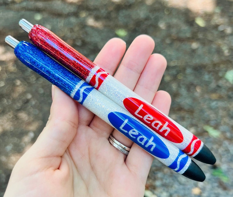 K&ouml;nnte beinhalten: Zwei personalisierte Glitzer-Stifte mit wei&szlig;en, roten und blauen Akzenten. Die Stifte sind mit einem Kreidendesign verziert und der Name "Leah" ist auf jedem Stift geschrieben.