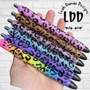 May include: A set of five glittery pens featuring a black leopard print pattern over various gradient colors including pink, gold, purple, blue, and yellow. The text Leah Danae Designs LDD is visible in the upper right corner of the image.
