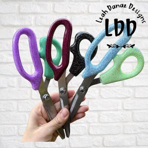 May include: A collection of five pairs of scissors with glittery, colorful handles in purple, teal, dark red, black, and light blue. The metal blades are joined by a central screw. The text Leah Danae Designs LDD is visible in the upper right corner.