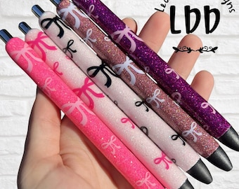 May include: Five glittery pens with black tips, each featuring a repeating bow pattern. The colors range from bright pink, light pink, white, rose gold, to deep purple. The text Leah Danae Designs LDD is visible in the upper right corner.
