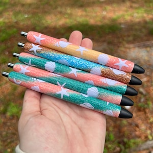 May include: Five pens with glitter and a seashell design. The pens are pink, blue, and green with white seashells and starfish.