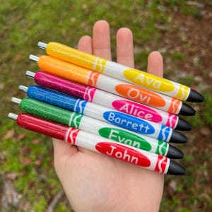 K&ouml;nnte beinhalten: Sieben Glitzer-Stifte mit Crayola-&auml;hnlichen Designs und darauf geschriebenen Namen. Die Stifte sind in den Farben Rot, Gr&uuml;n, Blau, Pink, Orange und Gelb.