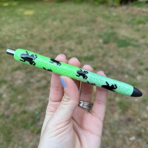May include: A green glitter pen with black frog silhouettes. The pen has a silver metal tip and a black tip. The pen is held in a hand with silver rings and blue nail polish.