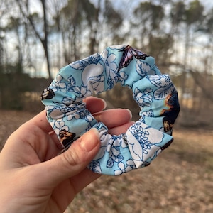 May include: A light blue scrunchie with a floral and butterfly pattern. The fabric features white flowers and butterflies with brown and pink wings. The scrunchie is a hair accessory.