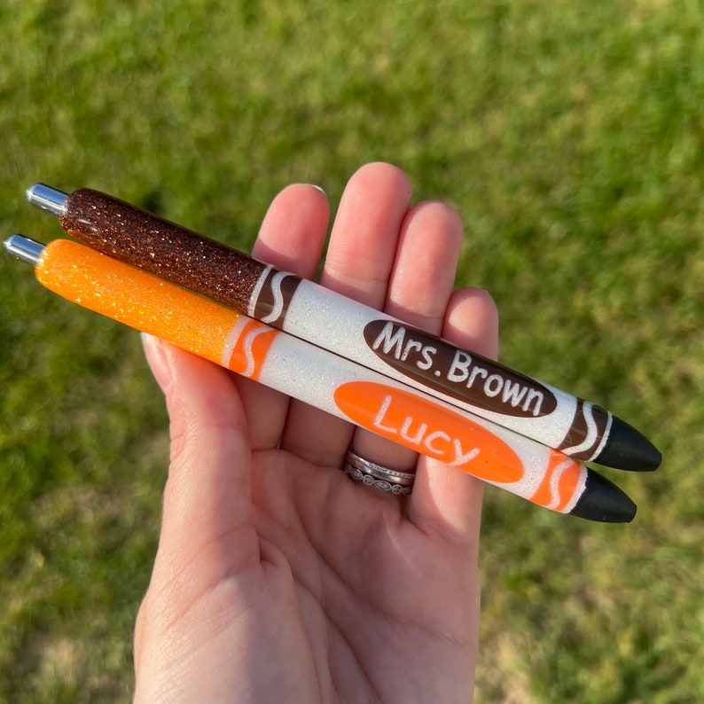 K&ouml;nnte beinhalten: Zwei Glitzer-Stifte in Form von Buntstiften. Der orange Stift sagt "Lucy" und der braune Stift sagt "Frau Brown".