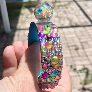 May include: A cuff bracelet featuring a bright blue interior and an exterior surface densely covered in multicolored, iridescent, faceted rhinestones. A clear glass sphere filled with tiny, colorful beads is attached to the top edge.