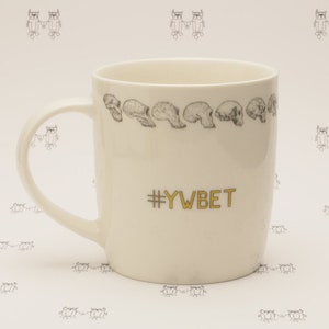Puede incluir: Taza de cerámica blanca con un dibujo en blanco y negro de cráneos alrededor del borde y el texto "#YWBET" en letras doradas.