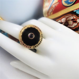 Può includere: Un anello d'oro con una pietra di onice nero incastonata in una lunetta circolare. L'anello è decorato con una fila di piccole pietre preziose rosa e nere attorno alla lunetta.