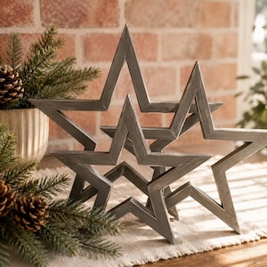 May include: Two five-pointed star decorations crafted from dark gray metal with a weathered finish. The stars are open-framed and layered, with one positioned slightly in front of the other, creating a dimensional display of geometric shapes.