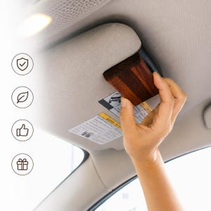 May include: A dark brown wooden clip attached to a light grey car sun visor. The clip features a visible wood grain texture and is positioned over a safety warning label on the visor. Four circular icons are arranged vertically on the left side.