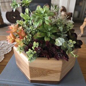 Op de afbeelding: Een houten, veelzijdige plantenbak gevuld met een verscheidenheid aan faux vetplanten in tinten groen, oranje en bordeaux. De plantenbak heeft een lichte houtnerf afwerking en staat op een blauw boek.