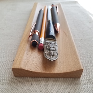 Può includere: Un portapenne in legno con un bordo curvo, che contiene una penna nera, una penna color oro rosa, una matita gialla e un aprilettere d'argento con un designo ornato.